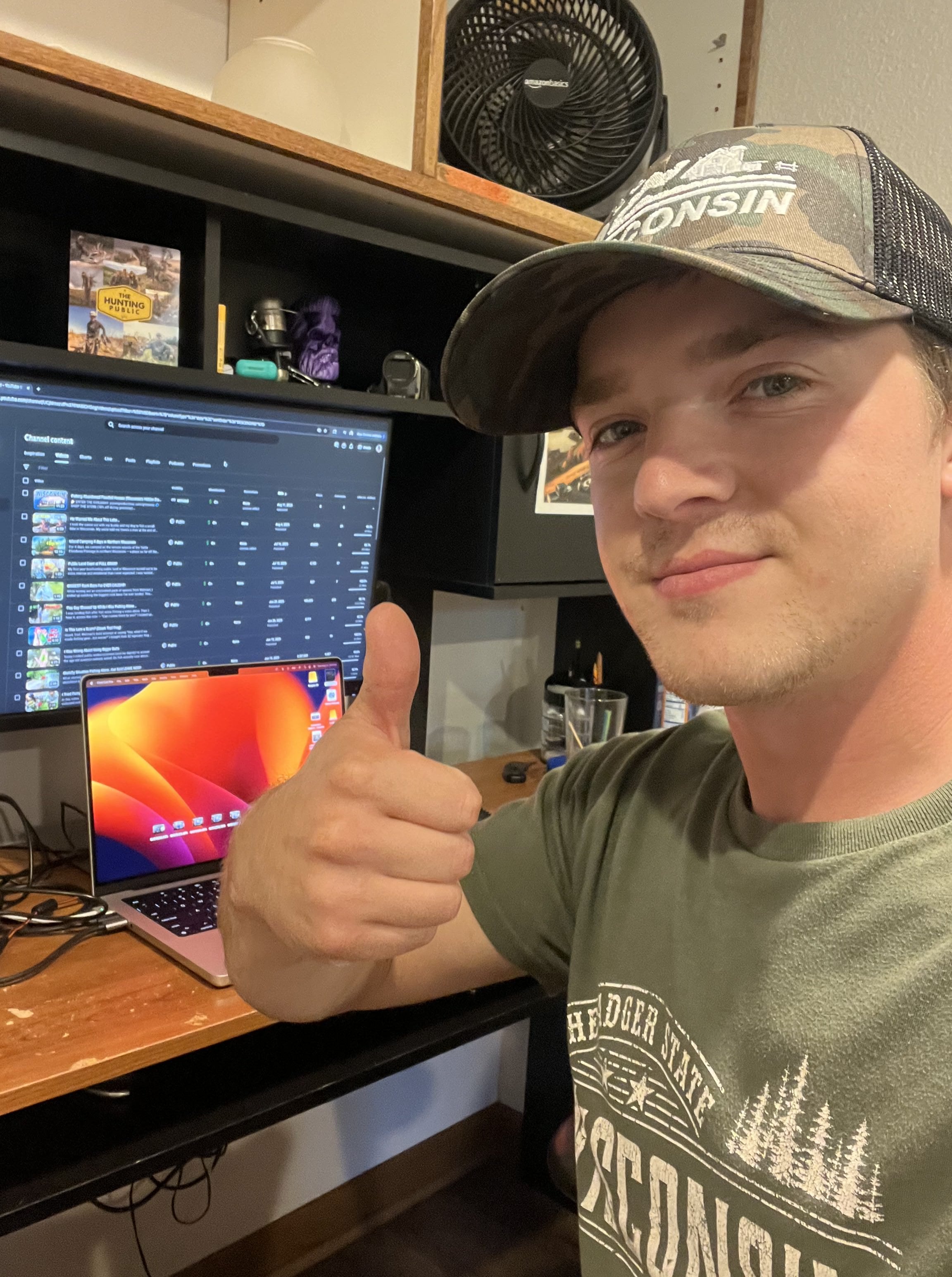 Person giving a thumbs up in front of a computer setup with multiple monitors while wearing a hat that says "Wisconsin."