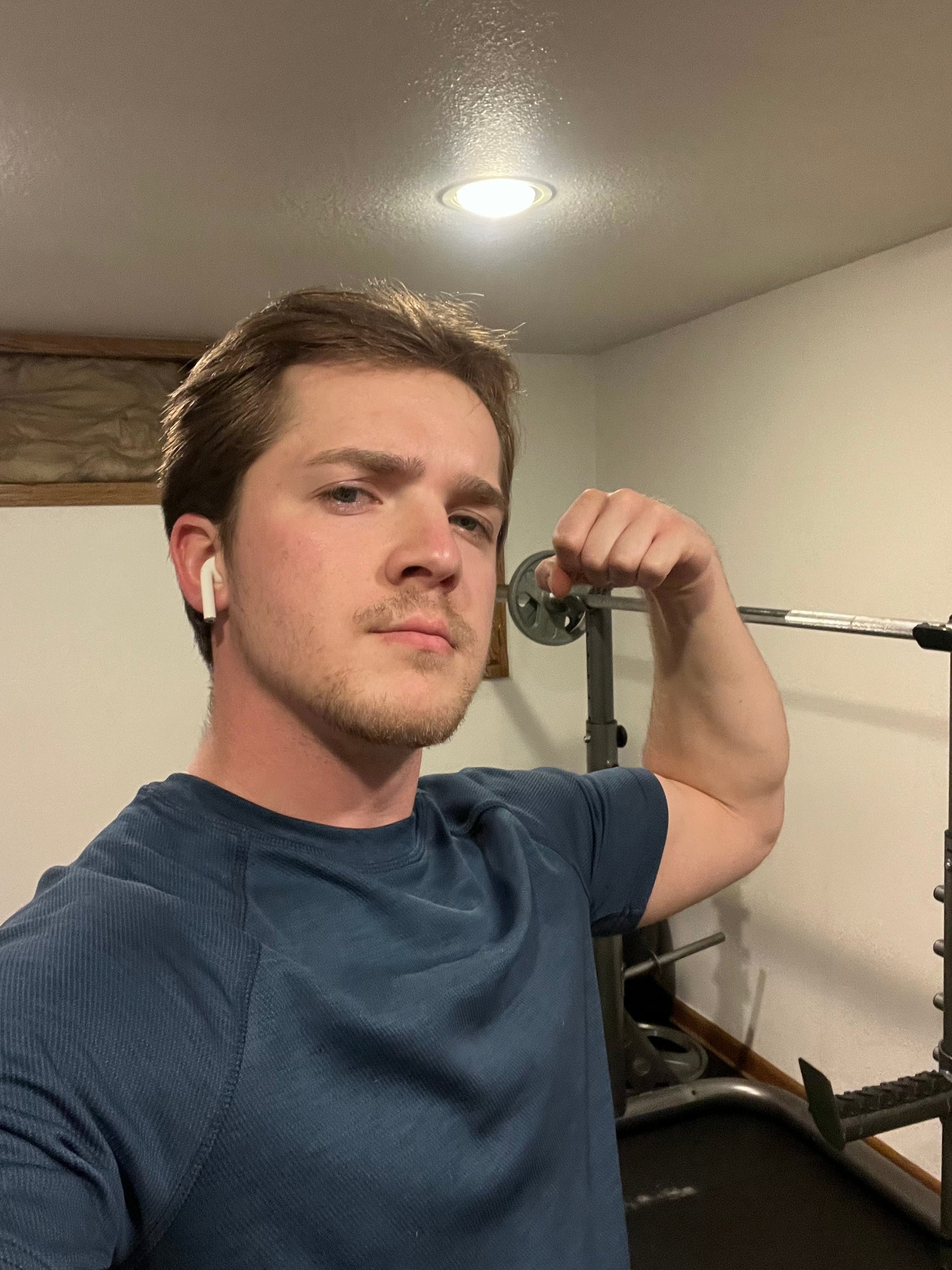 Man in blue shirt flexing next to weights and squat rack in basement with white walls. 