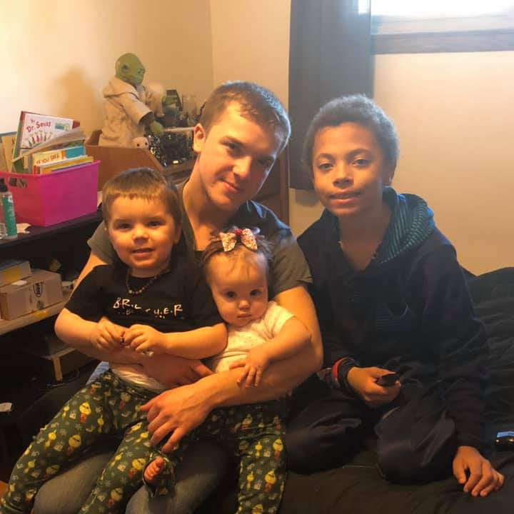 Three children sitting on a couch with a baby, in a casual indoor setting.