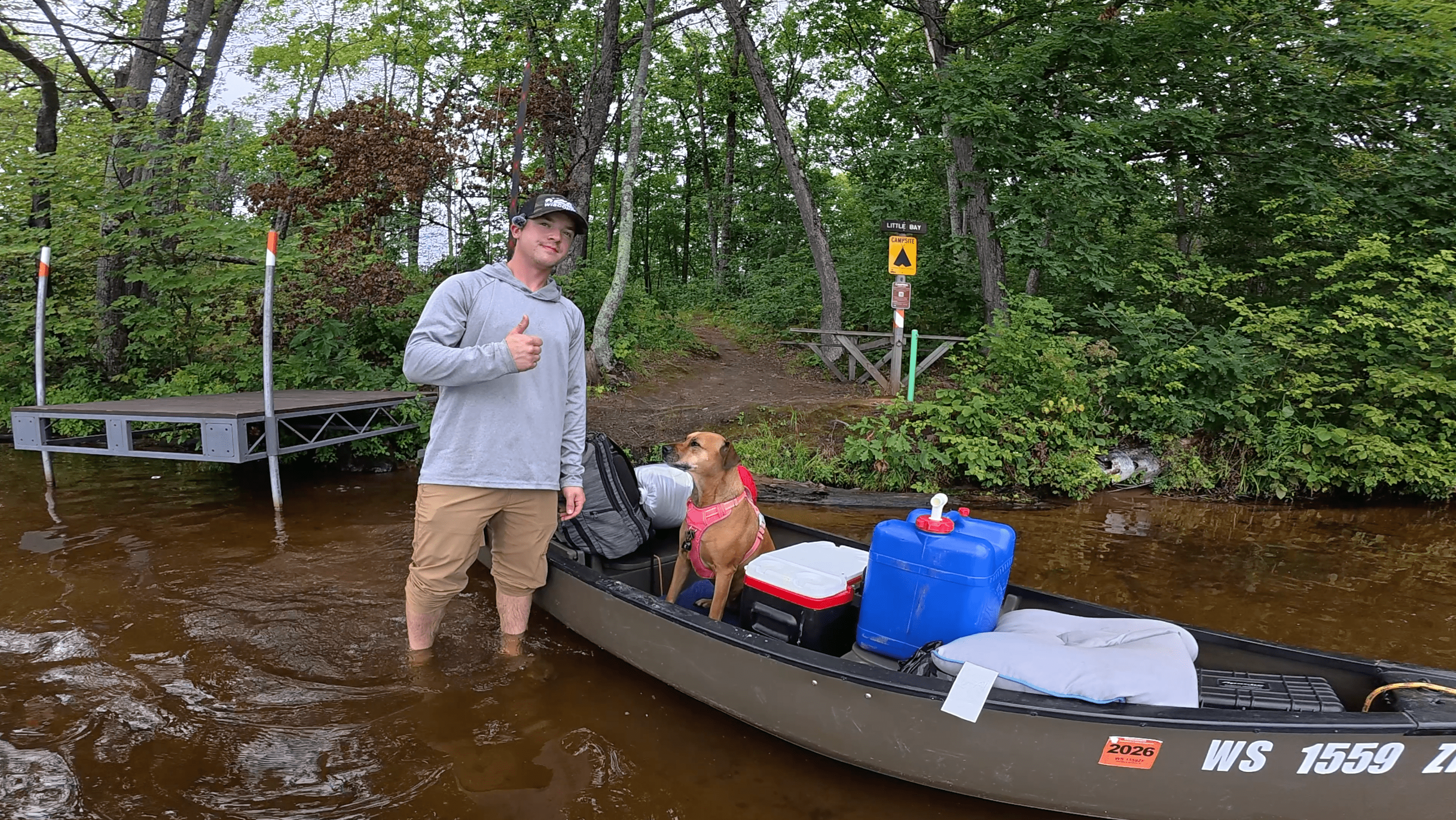 Man standing in water next to a canoe with a dog, surrounded by trees and a dock. Governor Thompson State Park in Wisconsin