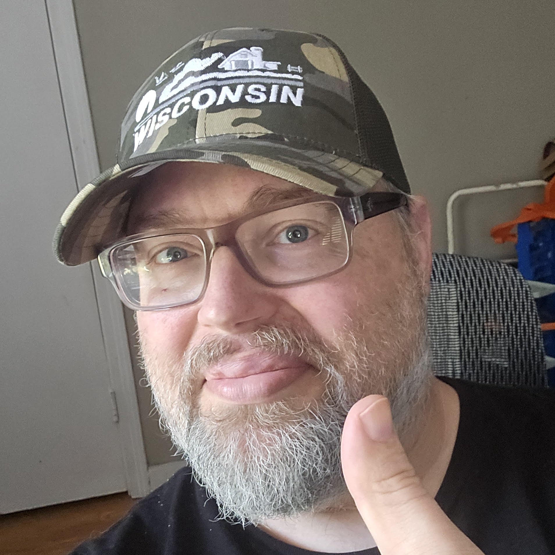 Man wearing a Wisconsin cap, giving a thumbs-up gesture indoors.